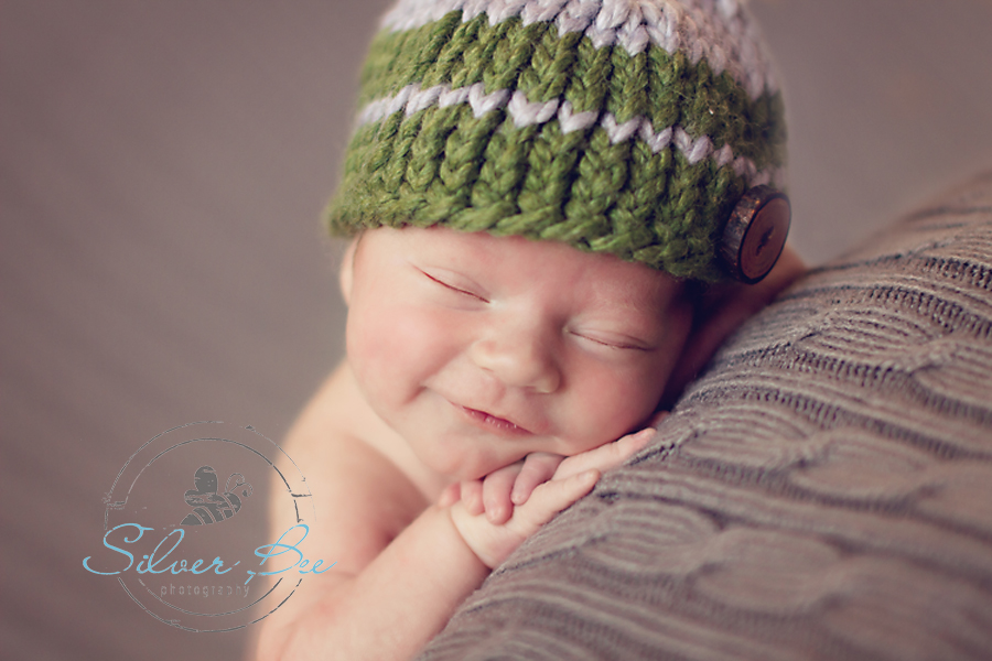 Austin newborn boy smiling in striped green hat and sleeping on gray cable blanket.