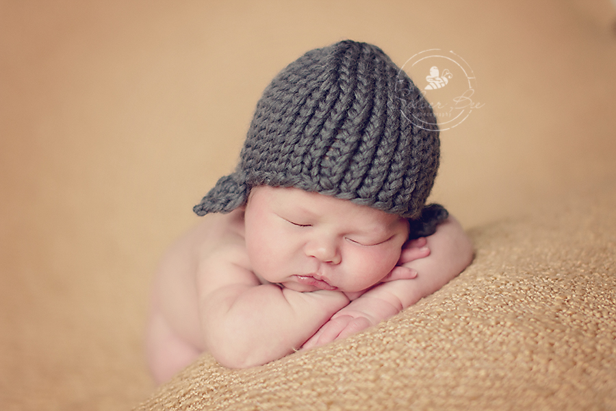 One week old newborn baby boy sleeping for newborn photo session on golden boucle blanket and dark gray aviator knitted hat in Austin Texas.